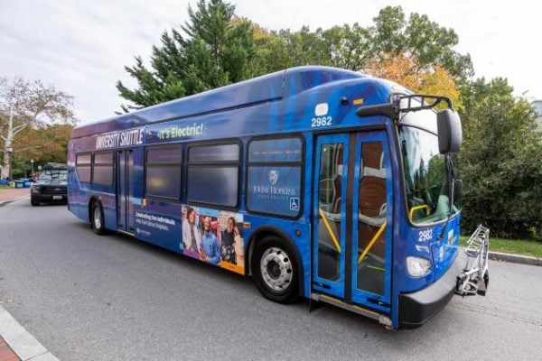A blue bus with "It's Electric" graphics coming around a curve on campus. 