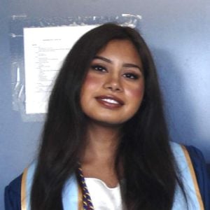 Reba Prabhakhar wearing graduate attire, standing against a blue wall. 