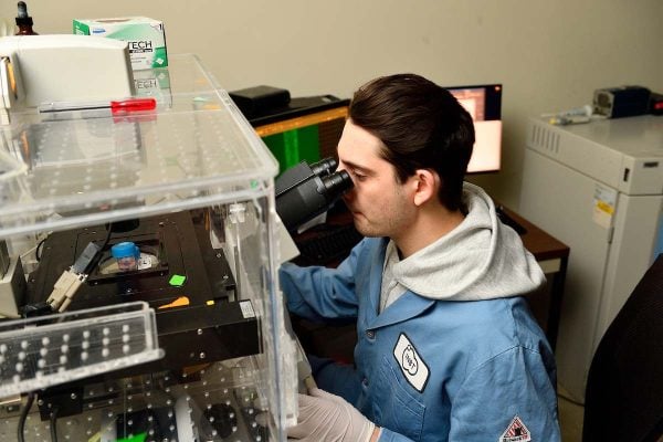 Alex Pessell looks through a microscope inside an enclosed imaging setup, with lab equipment and monitors nearby.