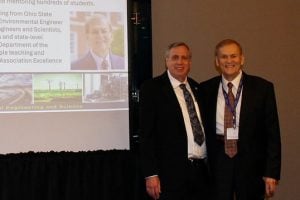 Hedy Alavi standing next to Jeffrey Greenfield in front of a screen showing a presentation