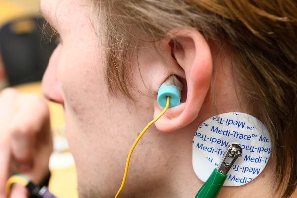 Close-up of a wearable in-ear EEG device with a foam earbud and electrode patch attached behind the ear for brain signal monitoring.